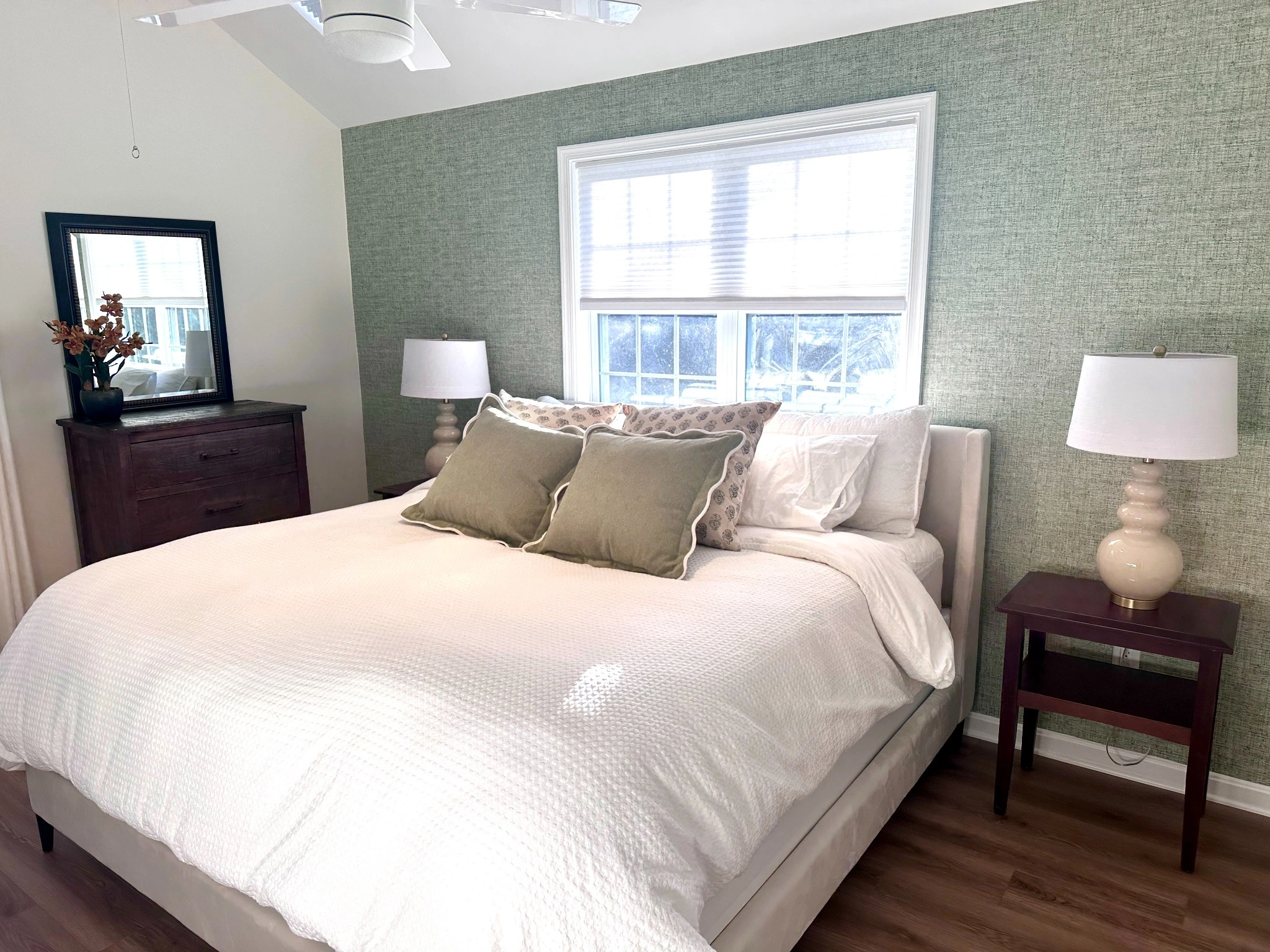Original bedroom with sage wallpaper, bright window light, loosely arranged bedding, and dark wood furniture